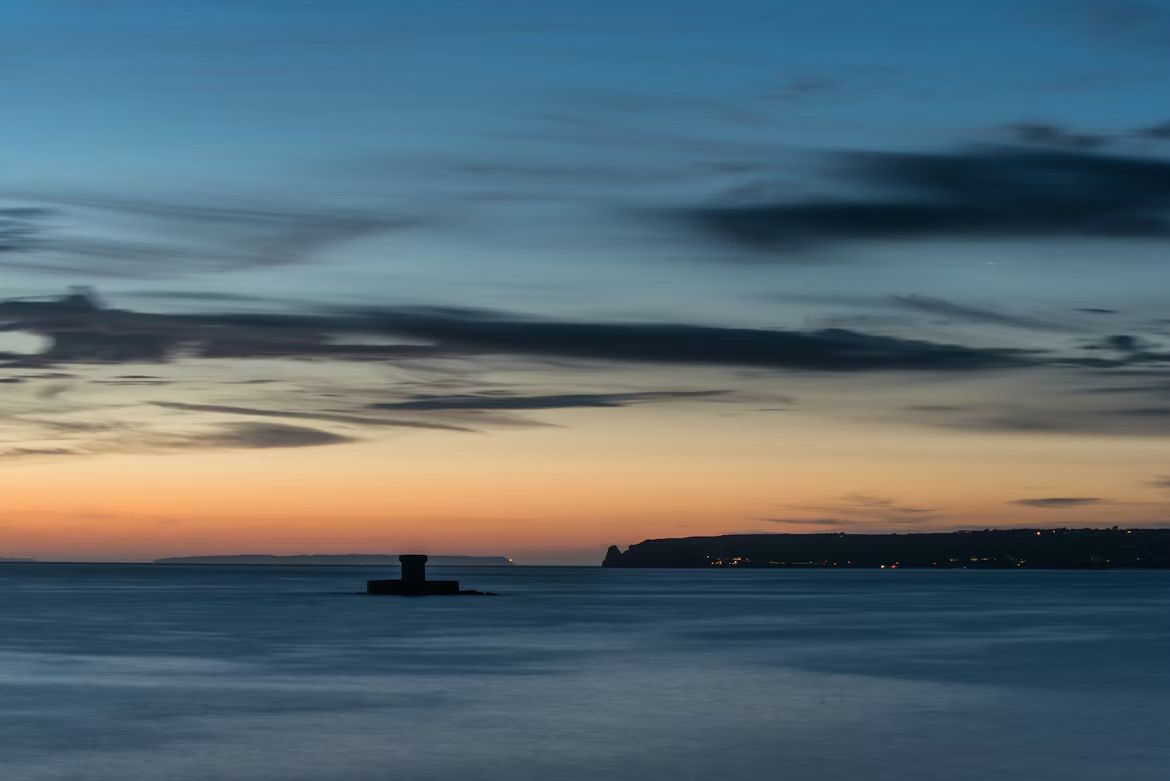 St Ouen's Bay at dusk seen from La Pulente, Jersey, Channel Islands, with La Rocco Tower silhouetted in the sea and lights along the coast.