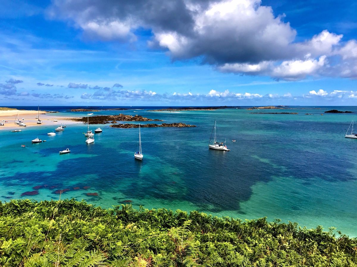Sailing and motor boats at anchor in clear turquoise water off a sandy beach on Herm, Channel Islands, seen from a bracken-covered hillside.