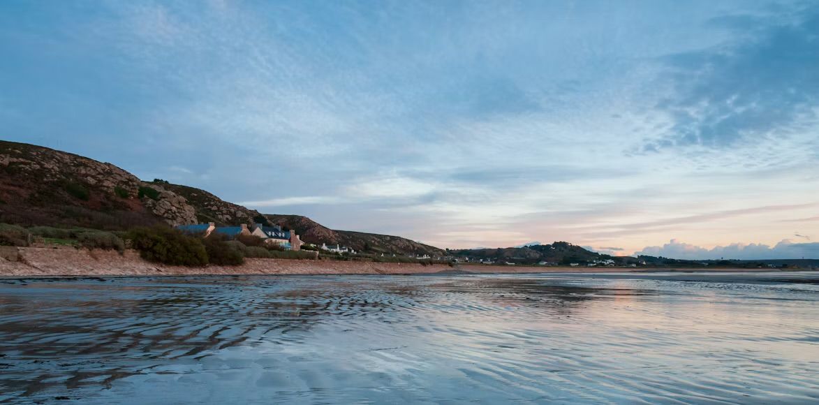 Wet sand at low tide below a rocky hillside and a row of cottages at L'Etacq, St Ouen's Bay, Jersey, Channel Islands, at dusk.