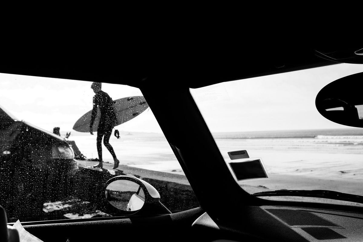 Black-and-white photo of a surfer carrying a board along a sea wall in Jersey, Channel Islands, seen through a car windscreen.
